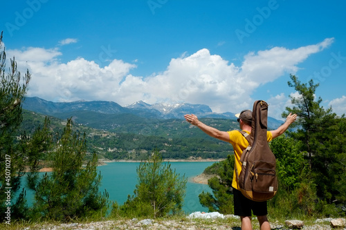 Young man wearing bright yellow t-shirt standing on top of the steep hill with the guitar. Male tourist enjoying the beautiful landscape view of mountain river and forest. Copy space, background.