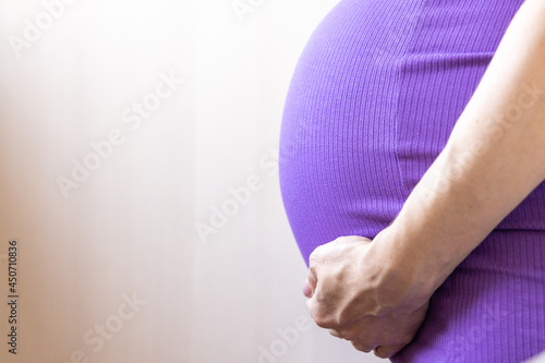 Unrecognizable Close up Pregnant women. Hands over tummy during last month of pregnancy. In violet color dress on light background. Hand on belly. Pregnancy, maternity, preparation concept.Copy space.