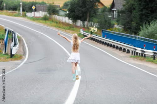Blonde girl wearing white clothes walking on a paved road with raised arms. Concept of local travel, summer in countryside, finding way.