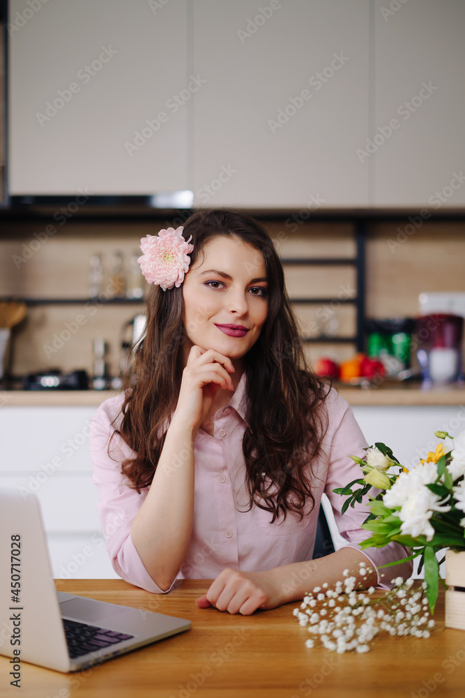 Young brunette woman with long wavy hair working on laptop with flowers on the desk with kitchen in background. Talented florist developing online sales getting ready for workshop.