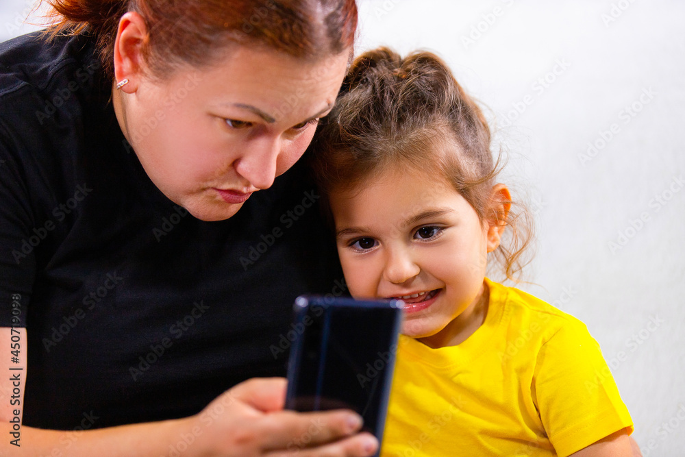 Close up photo of Mom and little girl sitting on the sofa and watching bad news headlines using mobile phone, they are addicted to the phone. Close up photo of Mom and little girl sitting on the sofa and watching bad news headlines using mobile phone, they are addicted to the phone.