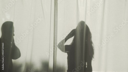 Silhouette of a long-haired woman and bold man standing behind curtains with their smartphones. Contrasting shadow in the bright sun. Summer mood. Young couple on vacation. People talking phone. Call.