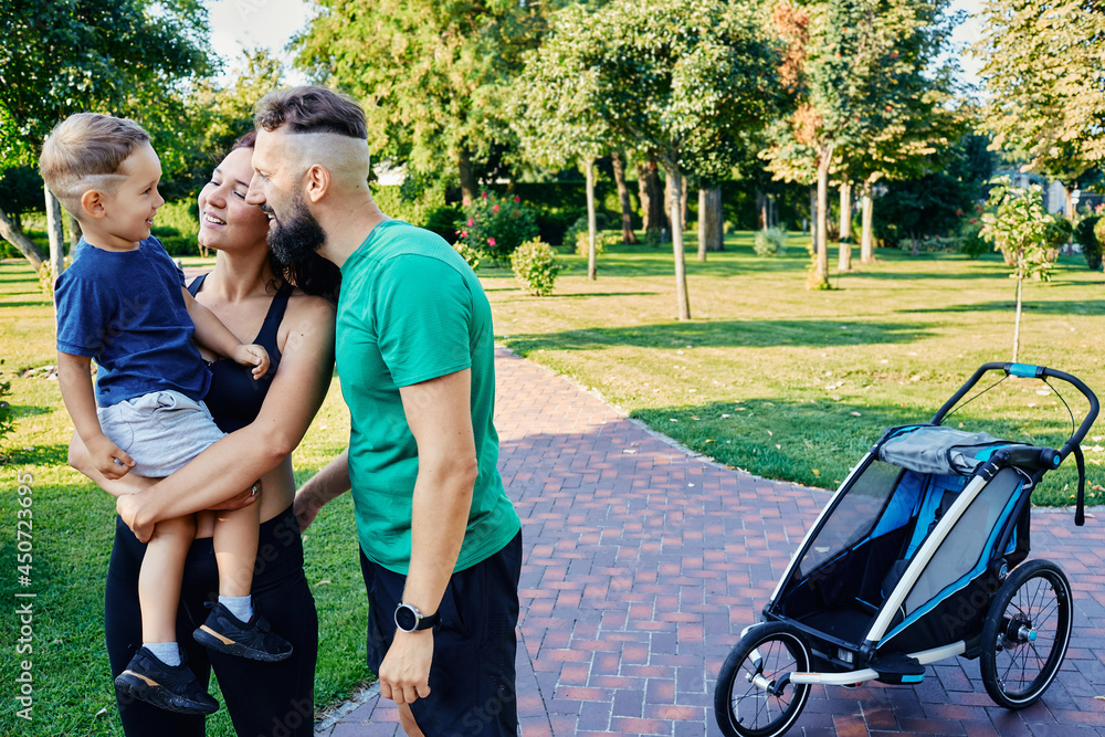 Active family with their son after a morning jog with a jogging stroller in a beautiful park. Sports for a whole family Active family with their son after a morning jog with a jogging stroller in a beautiful park. Sports for a whole family