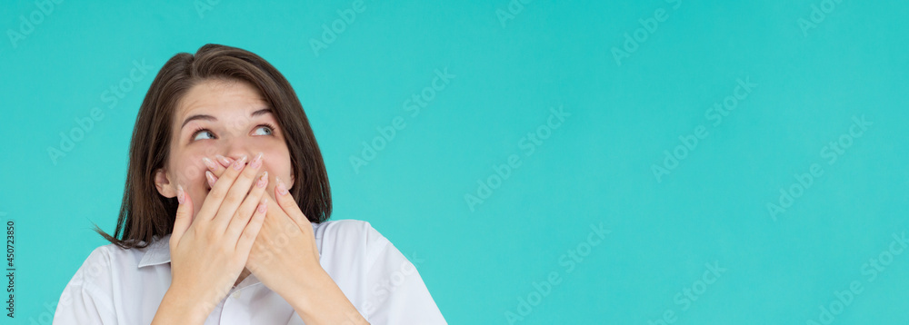 Shocked amazed laughing young brunette woman wearing casual basic white shirt standing covering mouth with hands isolated on pastel blue colour background studio portrait Shocked amazed laughing young brunette woman wearing casual basic white shirt standing covering mouth with hands isolated on pastel blue colour background studio portrait