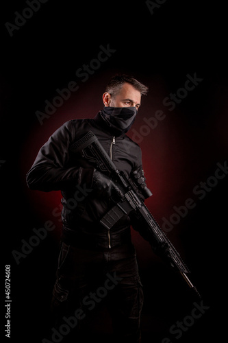 A man in dark clothes holds an automatic carbine in his hands. The face is covered with a mask, hands are in gloves. A figure with a weapon on a dark back with a red light spot.