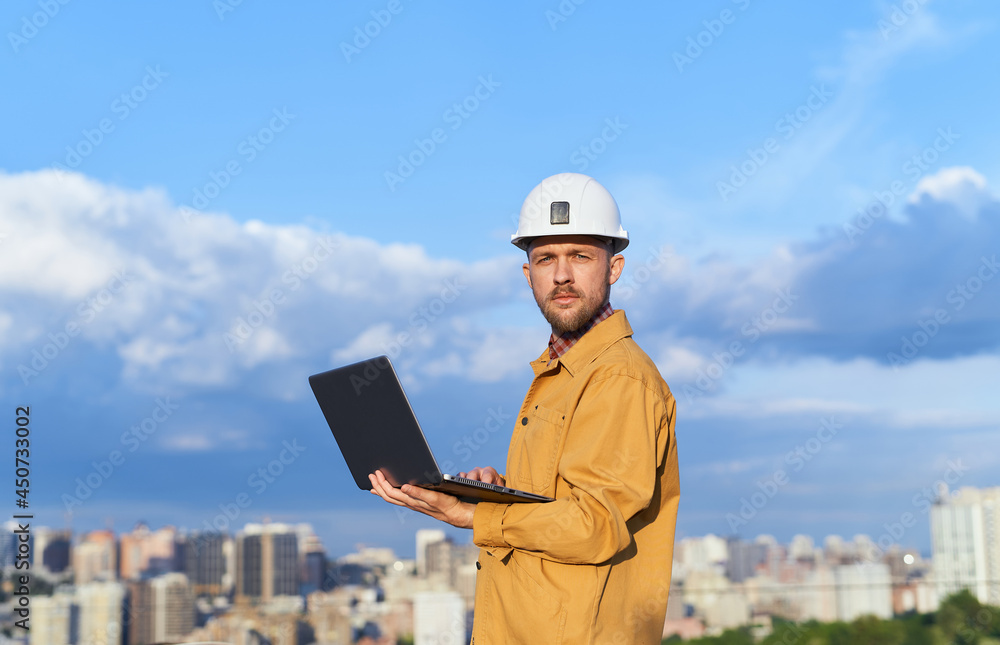 Engineering male on construction site holding laptop computer in his hand on urban city background and blue cloudy sky. Building inspector, contractor or supervisor concept. High quality photo Engineering male on construction site holding laptop computer in his hand on urban city background and blue cloudy sky. Building inspector, contractor or supervisor concept. High quality photo