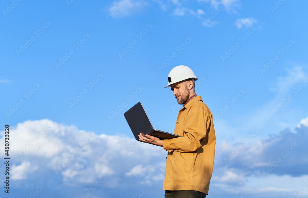 Attractive caucasian bearded supervisor, building contractor or engineer in white helmet and orange jacket using laptop at construction site. Sunny day with blue cloudy sky. High quality image Attractive caucasian bearded supervisor, building contractor or engineer in white helmet and orange jacket using laptop at construction site. Sunny day with blue cloudy sky. High quality image