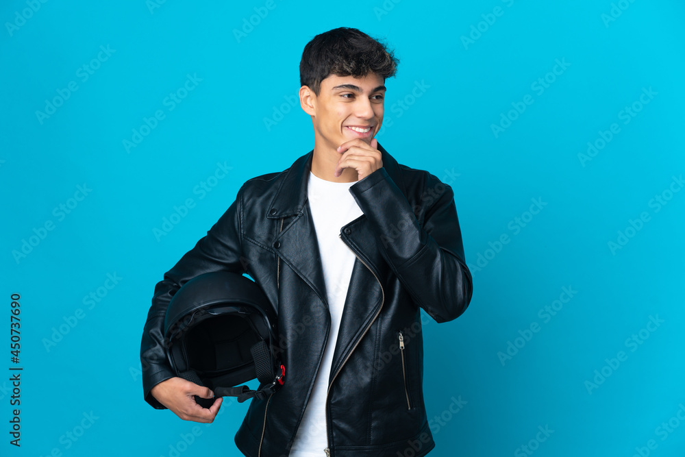 Young man with a motorcycle helmet over isolated blue background looking to the side and smiling Young man with a motorcycle helmet over isolated blue background looking to the side and smiling