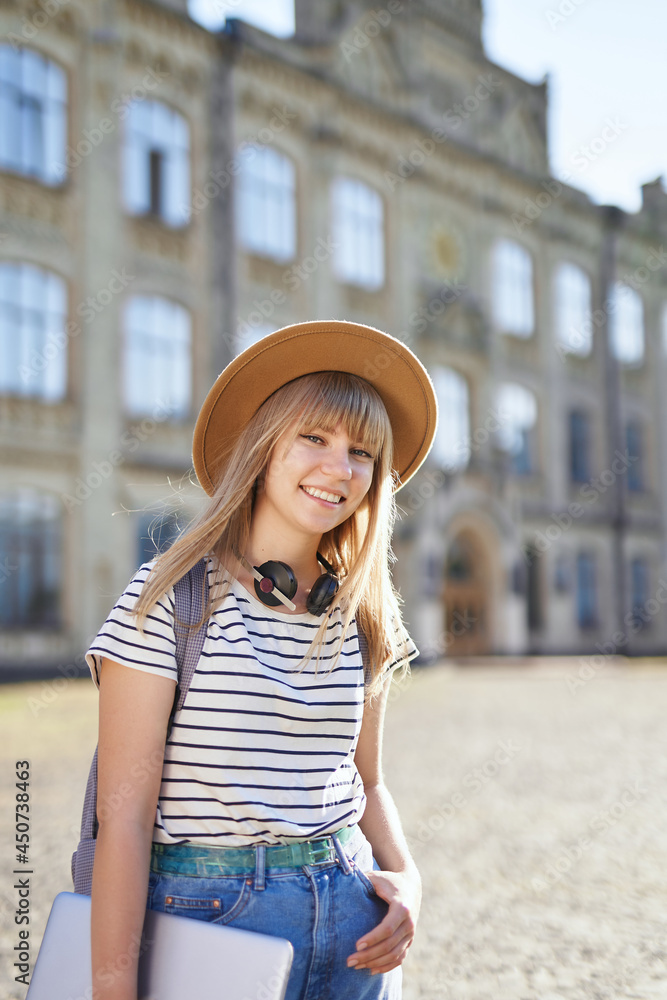 College girl, student portrait, studying abroad or educational concept. Cheerful smiling young blonde female European university or college student wearing brown hat at campus. High quality image