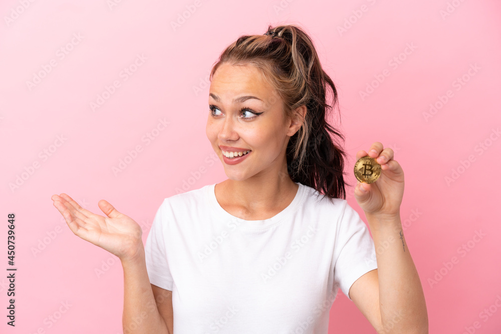 Teenager Russian girl holding a Bitcoin isolated on pink background with surprise expression while looking side Teenager Russian girl holding a Bitcoin isolated on pink background with surprise expression while looking side