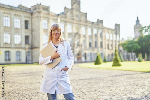 Intern or student doctor in university campus looking straight at camera. Education concept. Portrait of young blonde medical university student with laptop standing outdoor wearing white medical gown