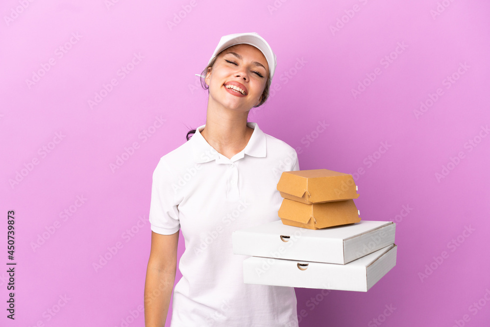 Pizza delivery Russian girl with work uniform picking up pizza boxes and burgers isolated on purple background laughing Pizza delivery Russian girl with work uniform picking up pizza boxes and burgers isolated on purple background laughing