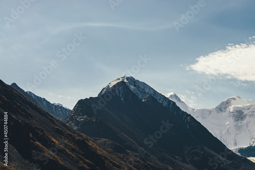 Atmospheric alpine landscape with high mountain silhouette with snow on peaked top under cirrus clouds. Dramatic mountain scenery with beautiful snow-covered pointy peak and high snowy mountain wall.