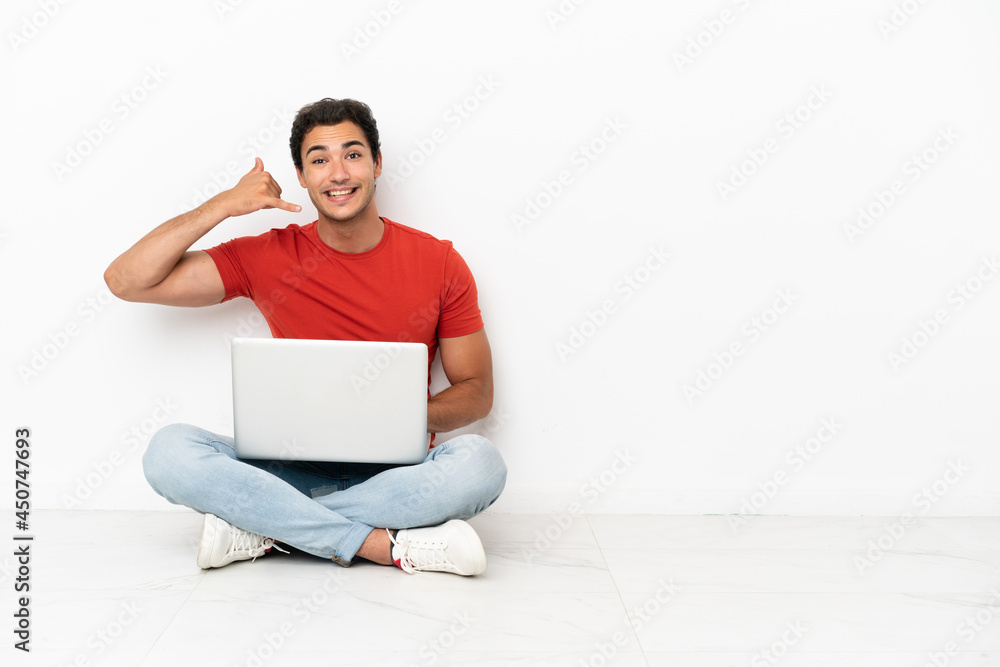 Caucasian handsome man with a laptop sitting on the floor making phone gesture. Call me back sign Caucasian handsome man with a laptop sitting on the floor making phone gesture. Call me back sign