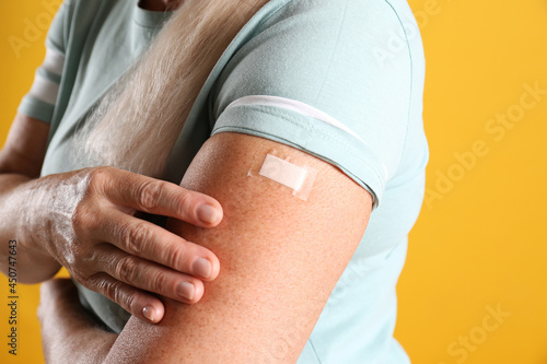 Mature woman showing arm with bandage after vaccination on yellow background, closeup
