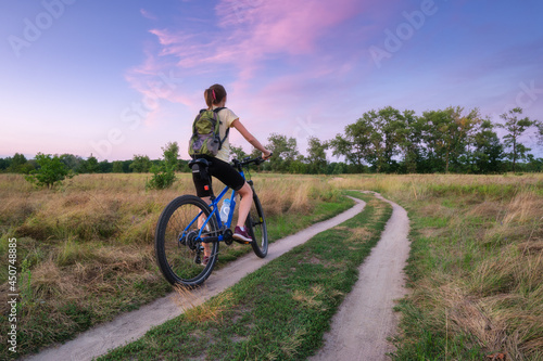 Woman is riding a mountain bike in cross country road at sunset in summer. Colorful landscape with sporty girl with backpack riding bicycle, field, dirt road, green grass, purple sky. Sport and travel