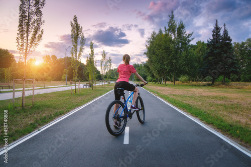 Fototapeta Naklejka Na Ścianę i Meble -  Woman riding a mountain bike on the asphalt road at sunset in summer. Colorful landscape with sporty girl riding a bicycle, road, green trees, grass and gold sunlite in park. Sport and travel. Cycle