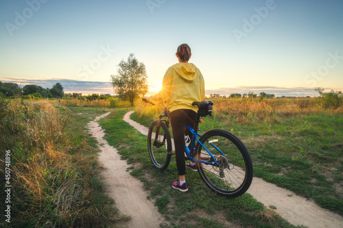 Fototapeta Naklejka Na Ścianę i Meble -  Woman is riding a mountain bike in cross country road at sunset in summer. Colorful landscape with sporty girl in yellow hoodie, bicycle, field, dirt road, green grass, trees, sky. Sport and travel