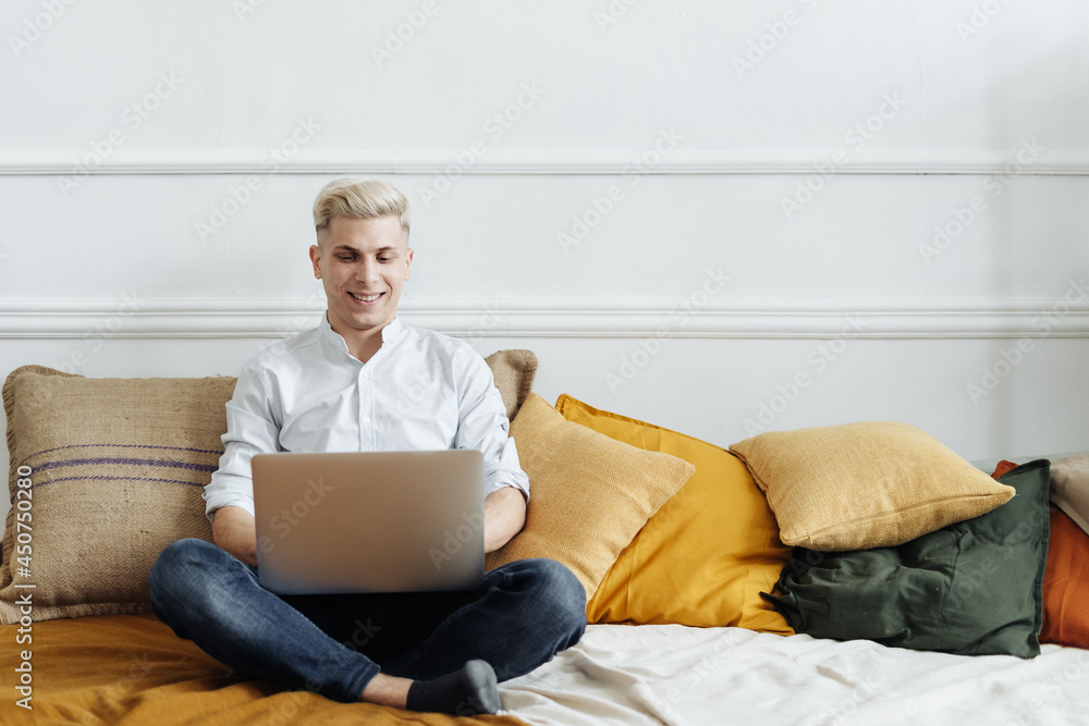 Young attractive smiling blonde guy is browsing at his laptop, sitting at home on the cozy sofa at home, wearing casual outfit Young attractive smiling blonde guy is browsing at his laptop, sitting at home on the cozy sofa at home, wearing casual outfit