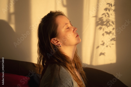 Young woman close eyes and relaxation in warm rays of sunset. Warm calm portrait of girl indoor