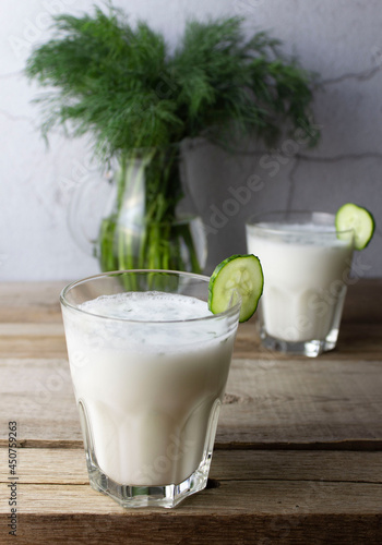 Ayran drink in a glass with a cucumber slice on a wooden table. Ayran with dill in a glass, a bunch of dill in a glass jug on the background