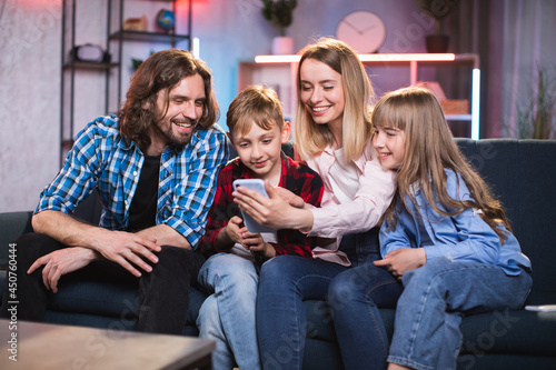 Two kids with their parents using cell phone for watching funny video or photos at home. Happy caucasian family in casual outfit sitting on couch and enjoying time together.