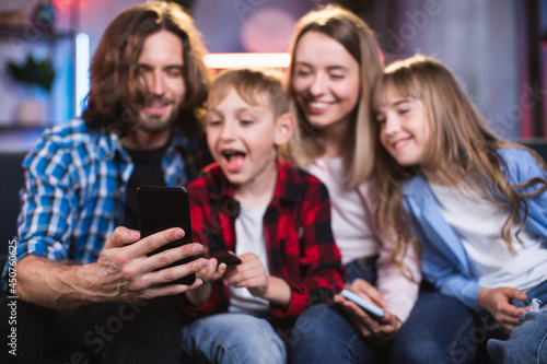 Playful kids and their young parents sitting in hugs on couch and taking selfie on modern cell phone. Concept of family  technology and leisure time.
