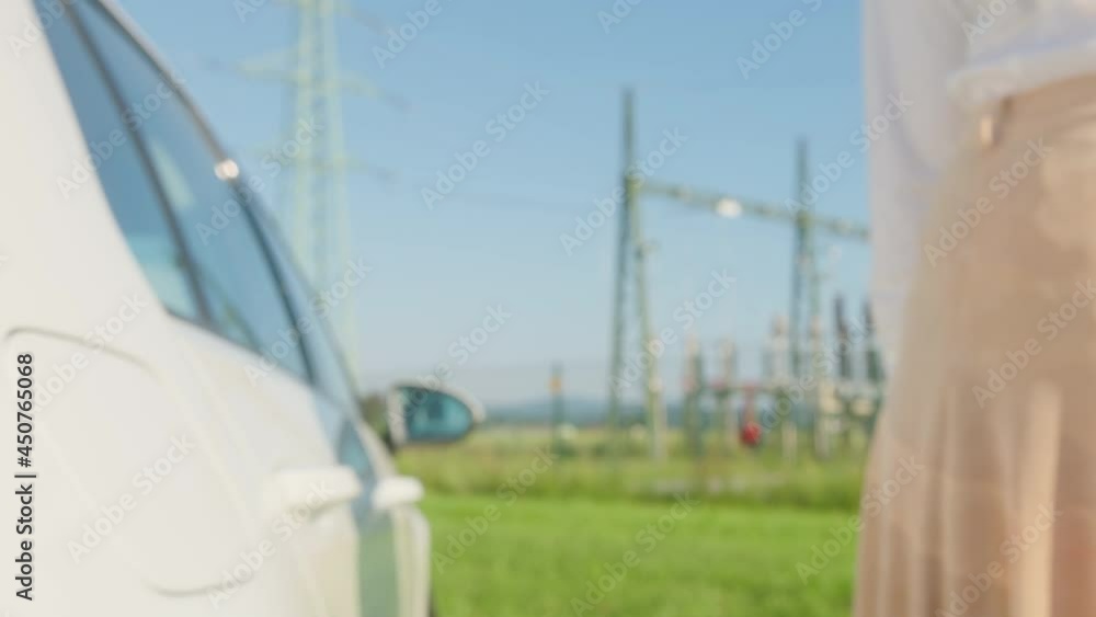 Extreme close up shot of a Caucasian female's hand holding an electric car charger and bringing it closer to the camera, in the background visible car and electric power plant Extreme close up shot of a Caucasian female's hand holding an electric car charger and bringing it closer to the camera, in the background visible car and electric power plant