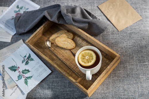 A white cup of tea with lemon, a long cupronickel spoon with a twisted handle and a saucer with three oatmeal cookies for breakfast on a wooden tray, a rag napkin on a gray sofa.