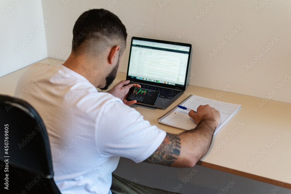 Close-up of a young Latino man with his back turned, working with cell phone and laptop and taking notes.