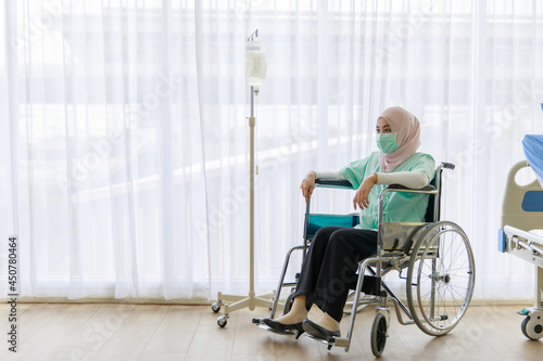 Portrait of ill Muslim wearing patient gown and face mask sitting on a wheelchair with saline fluid bag attach in hospital. Covid 19 pandemic concept