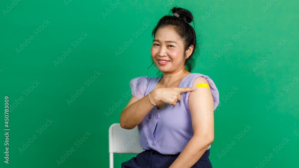 Studio shot of Asian middle aged woman sit smile pointing at yellow plaster bandage on arm after receive coronavirus covid 19 vaccination by doctor at clinic on green background Studio shot of Asian middle aged woman sit smile pointing at yellow plaster bandage on arm after receive coronavirus covid 19 vaccination by doctor at clinic on green background