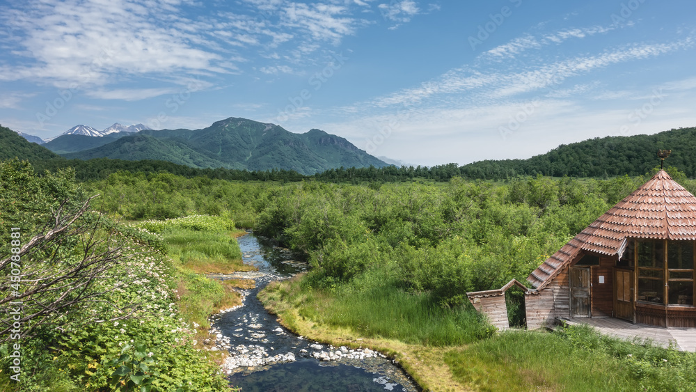 A stream with thermal water from a hot spring flows through the meadow. There is a wooden shed nearby. Lush green vegetation around. Mountains on the background of the sky and clouds. Kamchatka. A stream with thermal water from a hot spring flows through the meadow. There is a wooden shed nearby. Lush green vegetation around. Mountains on the background of the sky and clouds. Kamchatka.