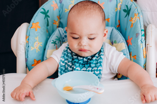 Serious baby sits at baby chair and looking to empty plate. Baby waiting for food in high chair
