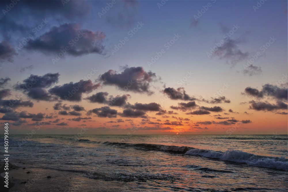 Early morning. The sun is rising above the horizon. There are orange lights in the sky, purple clouds. The waves of the Caribbean Sea are foaming on the sandy beach. Mexico 