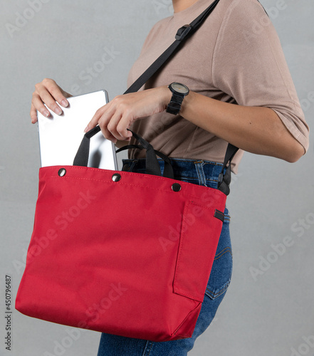Closeup studio shot of female model in brown long sleeve shirt putting silver notebook laptop computer into compartment pocket of trendy urban big red crossbody unisex handbag on gray background