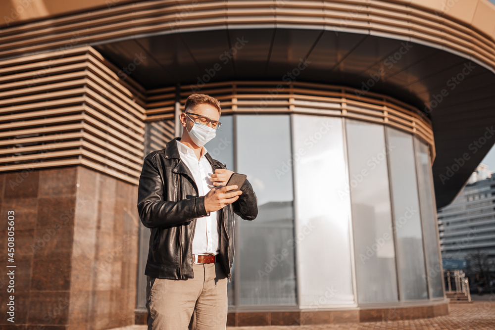 Handsome man in a protective mask on his face with glasses with a smartphone on the street of a big city. Businessman talking on the phone on urban background Handsome man in a protective mask on his face with glasses with a smartphone on the street of a big city. Businessman talking on the phone on urban background
