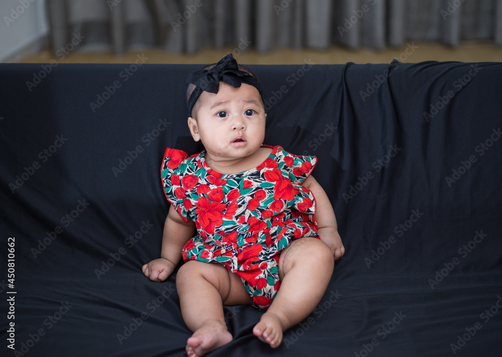 Fototapeta premium Little adorable baby girl sitting on black sofa alone. Asian 5 months old baby dressed in red dress with black headband sitting alone on sofa chair. 