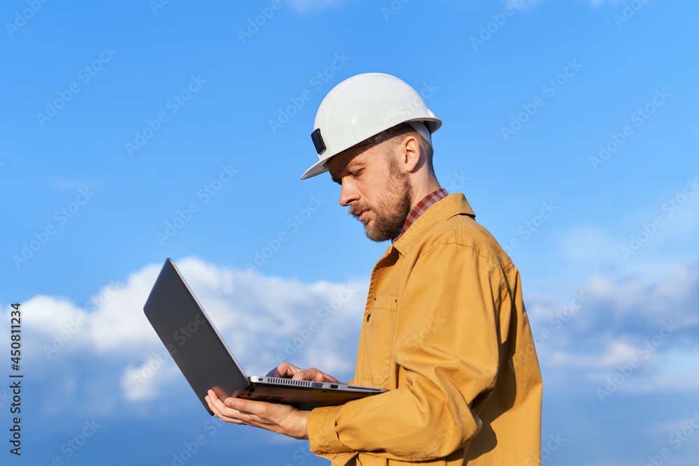 Building contractor, supervisor, instructor or construction engineer in white helmet and orange jacket using laptop at construction site. Sunny day with blue cloudy sky. High quality image Building contractor, supervisor, instructor or construction engineer in white helmet and orange jacket using laptop at construction site. Sunny day with blue cloudy sky. High quality image