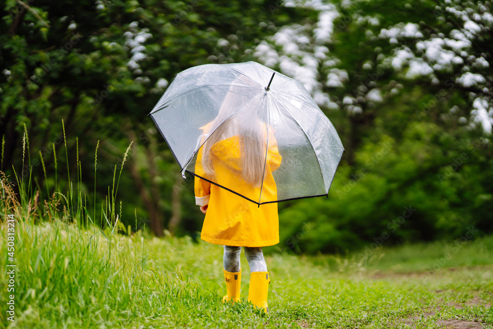 Little girl with transparent umbrella  playing in the rain on sunny autumn day. Beautiful girl  in a yellow cloak and rubber boots. Fall outdoor activity by rainy weather.
