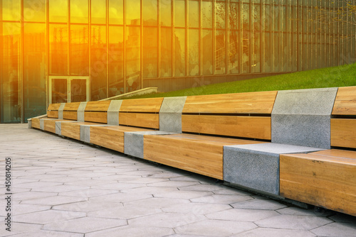 Wooden bench and tiled walkway. Office building for the work of company employees. Glass walls of a building with construction site reflection. Interior elements. Modern urban landscape.