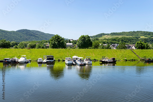 Yachts and motorboats moored at the marina on a calm river, forest covered hills are visible.