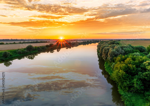 Scenic view at beautiful summer river sunset with reflection on water with green bushes, grass, golden sun rays, calm water ,deep cloudy sky and glow on a background, spring , evening landscape
