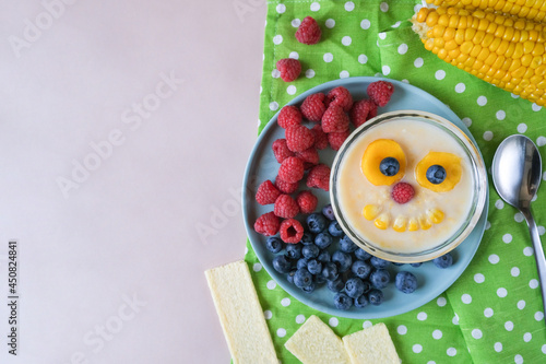 Fun food for kids - cute and smiling corn porridge monster with seasonal raspberry and blueberry decorations in the background. Inspiration for healthy baby cooking.
