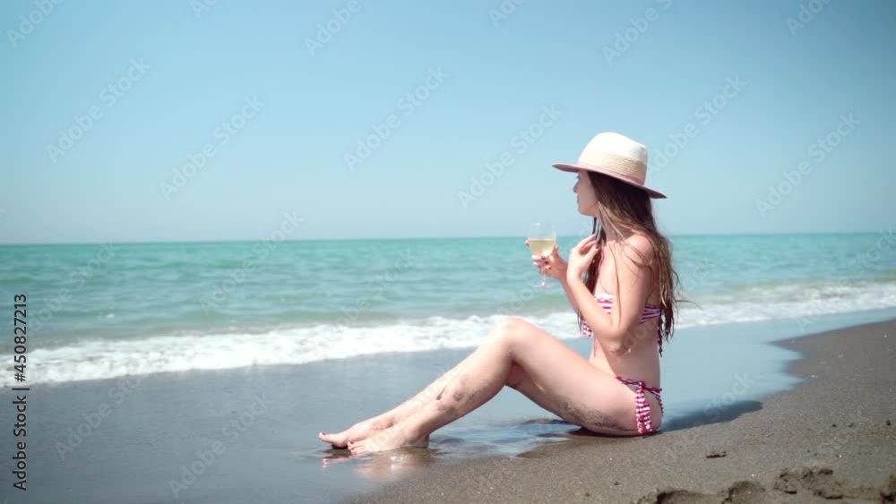 Pretty woman in sexy red and white swimsuit sitting at the sea beach being washed by salt water, pensive female with long brown hair and summer hat holding wineglass, drinking refreshing white wine Pretty woman in sexy red and white swimsuit sitting at the sea beach being washed by salt water, pensive female with long brown hair and summer hat holding wineglass, drinking refreshing white wine