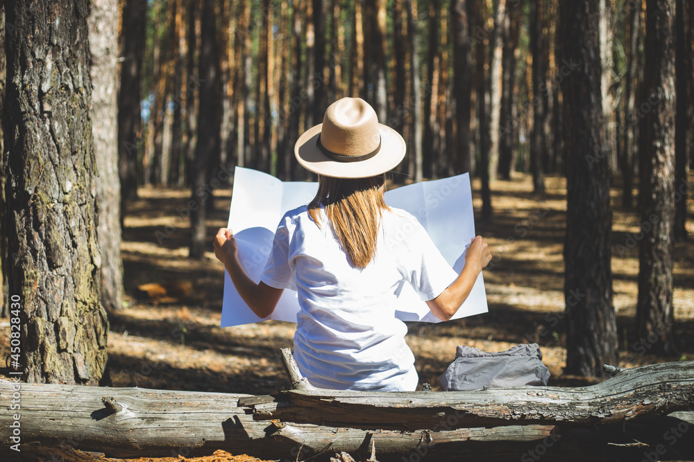 Young woman tourist in a hat and a t-shirt sits on a log and looks at a map during a halt in the forest. Back view
