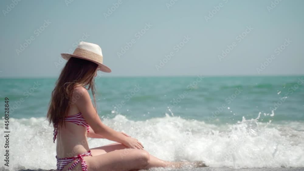 Warm rough ocean waves knocking down pretty smiling woman in summer hat sitting on the seashore. Happy caucasian woman in sexy bikini enjoying summer vacations, lying in water and playing with waves Warm rough ocean waves knocking down pretty smiling woman in summer hat sitting on the seashore. Happy caucasian woman in sexy bikini enjoying summer vacations, lying in water and playing with waves