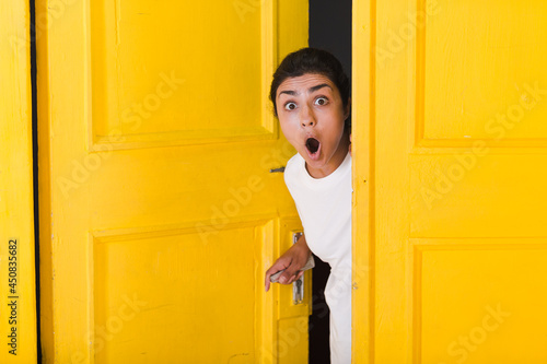 Фотография Young surprised indian woman peeking through yellow opened door