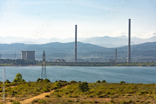 Two refrigeration towers and other chimneys from a power station next to a lake with some mountains on the background
