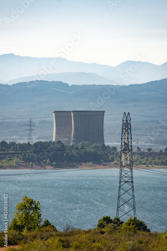 Two refrigeration towers from a power station next to a lake with some mountains on the background